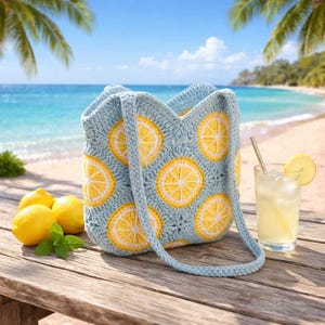 Op de afbeelding: Een lichtblauwe gehaakte draagtas met gele en witte citroen schijfjes patronen. De tas heeft een bijpassende band en staat op een houten tafel met citroenen, munt en een glas limonade. De achtergrond toont een strand.