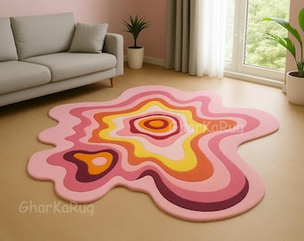 May include: An irregular, blob-shaped area rug featuring concentric, wavy layers in shades of light pink, bright yellow, orange, and deep burgundy. The rug displays a fluid, organic design with the text GharKaRug visible on the surface.