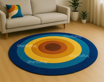 Può includere: Un tappeto circolare caratterizzato da anelli concentrici nelle tonalità blu scuro, ottanio, giallo, arancione e un centro marrone pieno. Il tappeto riporta il testo ripetuto "GharKaRug" sulla superficie in un carattere chiaro e semitrasparente.