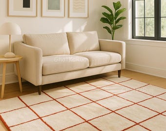 Puede incluir: Una alfombra rectangular con una base de color crema y un patrón de cuadrícula formado por líneas finas y texturizadas de color naranja óxido. La alfombra está sobre un suelo de madera clara frente a un sofá beige, con el texto GhatKaRug visible.