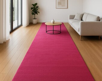 May include: A long, rectangular area rug in a solid, vibrant magenta pink color. The rug features a uniform, short-pile texture and is positioned centrally on a light-toned wooden floor, extending across the length of the room.