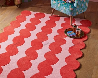 May include: A rectangular area rug featuring a repeating pattern of vertical wavy stripes in alternating shades of bright red and light pink. The rug has a distinct scalloped edge along its perimeter and includes the text GharkaRug printed on it.
