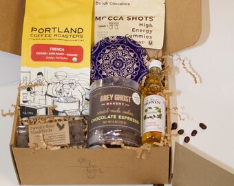 May include: A brown cardboard box filled with coffee, chocolate, and other treats. The box includes a bag of Portland Coffee Roasters French Roast coffee, a jar of Grey Ghost Bakery Chocolate Espresso cookies, a bottle of Monin Vanilla Syrup, a pack of Mocca Shots High Energy Gummies, and a blue and white patterned coaster. The box is surrounded by coffee beans.