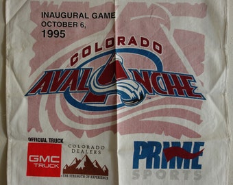 May include: A white cotton kitchen towel with a red, white, and blue Colorado Avalanche logo. The text "Inaugural Game October 6, 1995" is at the top of the towel. The text "Colorado Dealers" and "GMC Truck" are at the bottom of the towel.
