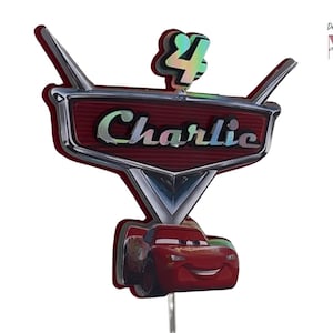 Peut inclure: Une décoration de gâteau personnalisée avec un emblème rouge en forme de voiture portant le nom Charlie en lettres irisées. Au-dessus se trouve le chiffre 4 avec une finition irisée. En dessous, un graphique de voiture de course rouge. Texte : Designed & Made by MPB Design.