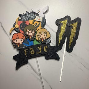 Peut inclure: Un topper de gâteau noir et or avec le numéro 11, un blason de Poudlard et le nom Faye. Le topper présente des personnages de la série Harry Potter.