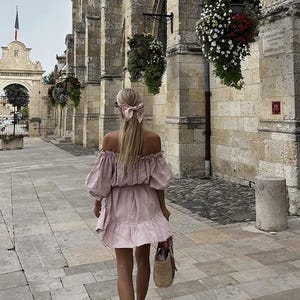 May include: A woman wearing a light pink off-the-shoulder dress with a ruffled hem walks down a stone pathway. She has blonde hair tied back with a floral patterned ribbon. She is carrying a woven straw tote bag.