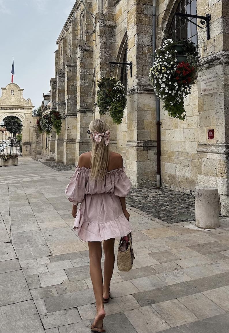 May include: A woman wearing a light pink off-the-shoulder dress with a ruffled hem walks down a stone pathway. She has blonde hair tied back with a floral patterned ribbon. She is carrying a woven straw tote bag.