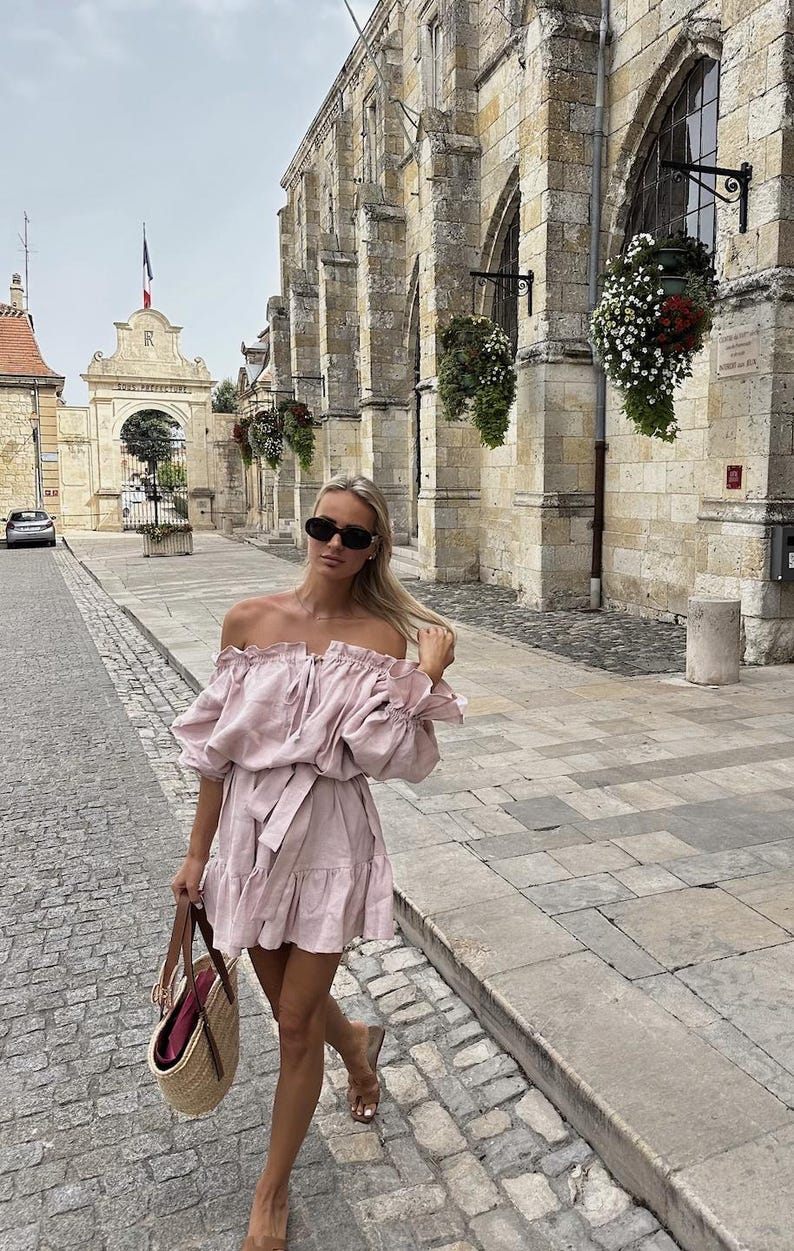 May include: A woman wearing a pink off-the-shoulder dress with a ruffled hem and a brown woven tote bag. She is walking on a cobblestone street in front of a stone building.