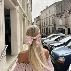 May include: A woman with long blonde hair wearing a pink off-the-shoulder top and a floral hair bow. She is walking down a street with cars parked on the side.