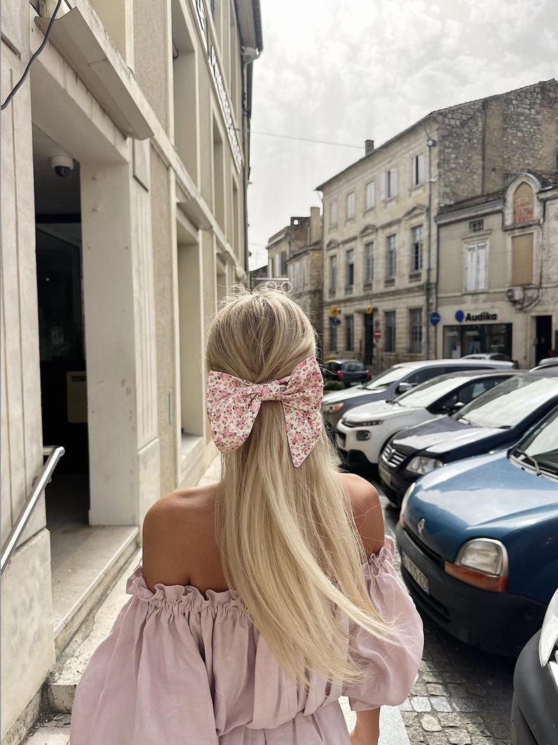 May include: A woman with long blonde hair wearing a pink off-the-shoulder top and a floral hair bow. She is walking down a street with cars parked on the side.