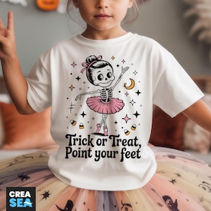 May include: White t-shirt featuring a skeleton ballerina in a pink tutu, with stars, a crescent moon, and candy corn accents. The text "Trick or Treat, Point your feet" is printed below the graphic. The shirt is part of a Halloween-themed collection.