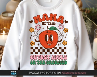 May include: A white crewneck sweatshirt featuring a graphic print of a cartoon apple with a face and arms making peace signs. The text reads MAMA OF THE CUTEST APPLE IN THE ORCHARD, surrounded by flowers, hearts, and a brown and white checkered pattern.