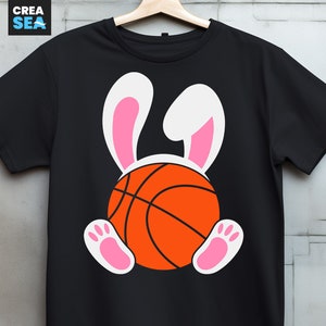 May include: Black t-shirt featuring a basketball with bunny ears and paws. The basketball is orange with black lines, and the bunny ears and paws are white and pink. The shirt is hanging on a wooden hanger.