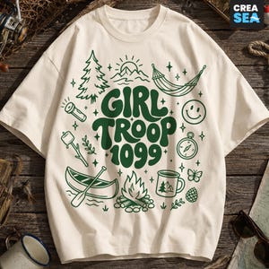 May include: A cream-coloured short-sleeve t-shirt featuring dark green graphic illustrations of camping gear, including a canoe, campfire, hammock, and compass, with the text "GIRL TROOP 1099" printed in a bold, wavy font in the centre.