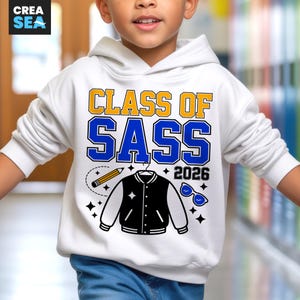 May include: White hoodie featuring the text "CLASS OF SASS 2026" in yellow and blue. The design includes a pencil, a varsity jacket, and sunglasses. The hoodie is worn by a child.