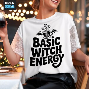 Könnte beinhalten: Weißes T-Shirt mit schwarzem Text "BASIC WITCH ENERGY". Das Shirt hat eine Grafik einer Kaffeetasse mit Fledermausflügeln und einem Pentagramm. Die Ärmel sind aus Spitze.