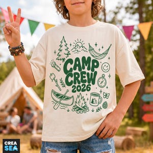Op de afbeelding: Een crèmekleurig t-shirt met een donkergroene grafische print met de tekst CAMP CREW 2026. Het ontwerp bevat illustraties van een dennenboom, zon, hangmat, zaklamp, marshmallow aan een stok, kano, kampvuur, mok en een smileygezicht.