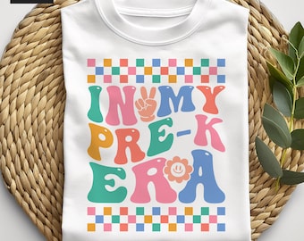 May include: A white t-shirt featuring a colorful graphic with the text "IN MY PRE-K ERA". The design includes a peace sign hand gesture, a smiley face flower, and rows of multicolored checkered squares in pink, blue, green, and orange.