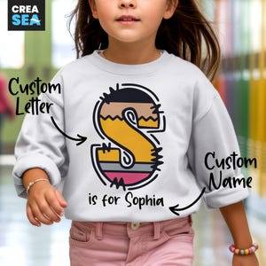 May include: White sweatshirt featuring a large, colorful letter "S" with a black outline. The letter is filled with shades of yellow, brown, and pink. The text "is for Sophia" is below the letter. The words "Custom Letter" and "Custom Name" are also visible.