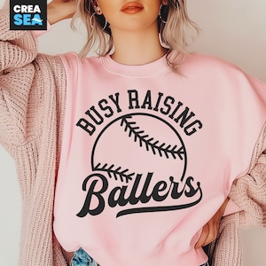 May include: Pink sweatshirt with a black baseball graphic and the text "Busy Raising Ballers".