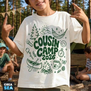 May include: An off-white short-sleeve t-shirt featuring a dark green graphic print with the text COUSIN CAMP 2026. The design includes illustrations of a pine tree, sun, hammock, flashlight, marshmallow on a stick, canoe, campfire, mug, and compass.