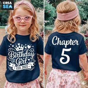 Pode incluir: Camiseta azul marinho com o texto "Birthday Girl EST. 2021" e um desenho de coroa. A parte de trás da camiseta diz "Chapter 5". A menina está usando uma faixa de cabelo e óculos de sol rosa, e uma saia rosa com estampa de borboletas.