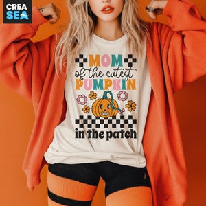 Puede incluir: Camiseta color crema con el texto "MOM of the cutest PUMPKIN in the patch" en una fuente retro. El diseño incluye una calabaza sonriente, flores y patrones de cuadros. La camiseta se combina con pantalones cortos negros y leggings a rayas naranjas.