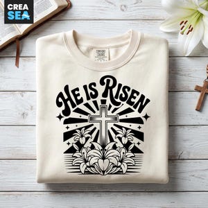 May include: Cream-colored sweatshirt with the text "He Is Risen" in a retro font, featuring a cross, lilies, and starburst design in black. The sweatshirt has a crew neck and a Comfort Colors tag.