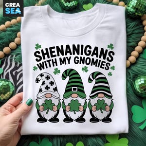 May include: White t-shirt with the text "SHENANIGANS WITH MY GNOMIES" and three gnome illustrations. Each gnome holds a green shamrock. The gnomes have different hat patterns: black and white stars, green stripes, and black and white stripes.