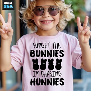 Könnte beinhalten: Hellrosa T-Shirt mit dem Text "FORGET THE BUNNIES I'M CHASING HUNNIES" in Schwarz. Das Shirt zeigt vier schwarze Hasen-Grafiken. Ein Kind mit Sonnenbrille und weißem Hut zeigt das Friedenszeichen.