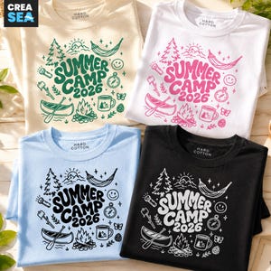 Puede incluir: Cuatro camisetas en crema, blanco, azul claro y negro, cada una con un gráfico de "Summer Camp 2026". El diseño incluye árboles, una hamaca, una brújula, una canoa, una fogata y una taza. Las camisetas son de "Hard Cotton".