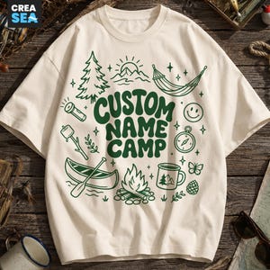 May include: A cream-coloured short-sleeve t-shirt featuring a dark green graphic print with the text CUSTOM NAME CAMP. The design includes illustrations of a pine tree, sun, hammock, smiley face, compass, marshmallow on a stick, canoe, and campfire.