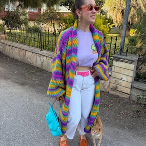 Op de afbeelding: Een vrouw draagt een kleurrijk gestreept gebreid vest met een lichtpaars t-shirt, een witte broek en oranje schoenen. Ze houdt een turquoise tas vast en wordt vergezeld door een rode kat. Het vest heeft een regenboog aan kleuren.