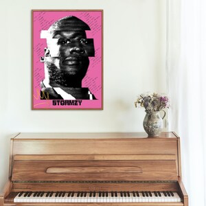 May include: A framed art print featuring a black and white halftone portrait of a person against a pink background. The word "STORMZY" is printed in black, with handwritten text and a gold crown graphic. The artwork is displayed above a wooden piano.