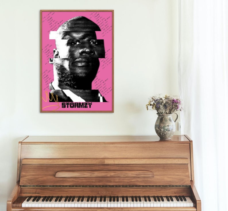 May include: A framed art print featuring a black and white halftone portrait of a person against a pink background. The word "STORMZY" is printed in black, with handwritten text and a gold crown graphic. The artwork is displayed above a wooden piano.