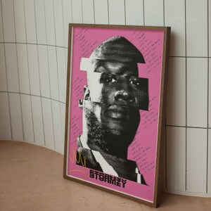 May include: A framed poster featuring a black and white portrait of a man with a pink background. The image has a fragmented design with text and the name "STORMZY" at the bottom. The frame is a warm brown color.