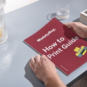 May include: A person holding a red "How to Print Guide" booklet with the text "MadebyReg." and a printer graphic. A glass of water and a decorative glass container with a yellow object inside are also visible on a gray surface.
