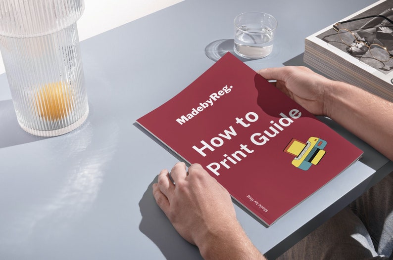 May include: A person holding a red "How to Print Guide" booklet with the text "MadebyReg." and a printer graphic. A glass of water and a decorative glass container with a yellow object inside are also visible on a gray surface.