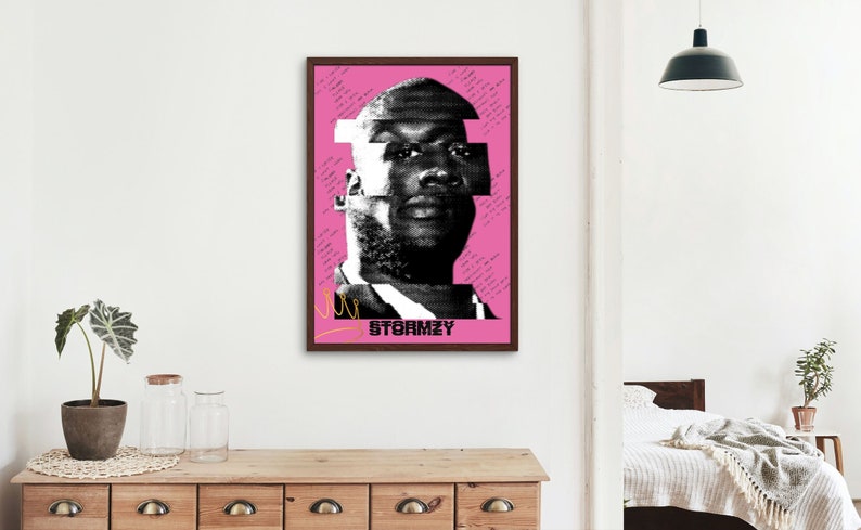 May include: A framed art print featuring a black and white portrait of Stormzy on a pink background. The image has a glitch art style with text elements and a yellow line. The print is in a dark brown frame.