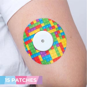 Puede incluir: Un parche redondo y colorido con un patrón de ladrillos estilo Lego en rojo, amarillo, verde, azul y morado. Un dispositivo circular blanco está centrado en el parche. El parche está adherido al brazo de una persona.