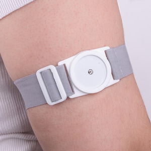 Op de afbeelding: Een grijze elastische armband met een witte plastic sluiting en een cirkelvormige witte sensor. De band is om een arm bevestigd, met de sensor op de bovenarm. De band heeft een gesp voor verstelling.