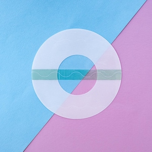 Pode incluir: Um objeto circular translúcido com um buraco central, dividido por uma linha diagonal. Uma faixa turquesa com um desenho ondulado cruza o centro. O fundo é dividido diagonalmente, com secções azuis e rosa.