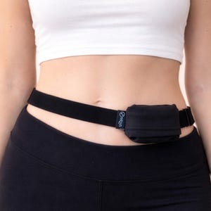 Op de afbeelding: Zwarte tailleband met een klein, vierkant zakje. De riem wordt om de taille gedragen en is gemaakt van een rekbaar materiaal. Het zakje is zwart en heeft een klein logo aan de zijkant. De persoon draagt een witte crop top en zwarte leggings.