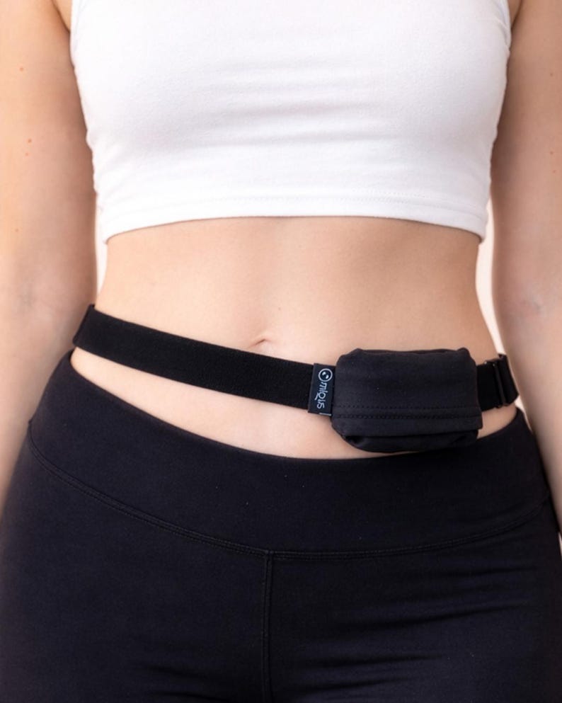 May include: Black waist belt with a small, square pouch. The belt is worn around the waist and is made of a stretchy material. The pouch is black and has a small logo on the side. The person is wearing a white crop top and black leggings.
