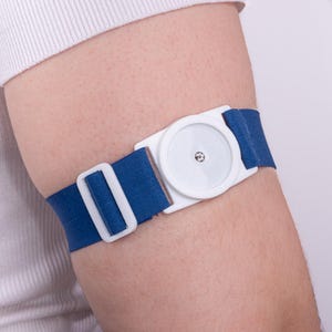 Op de afbeelding: Een blauw-witte medische tourniquet is om een arm gewikkeld. De tourniquet heeft een witte plastic gesp en een blauwe stoffen band. De gesp heeft een cirkelvormig ontwerp in het midden.