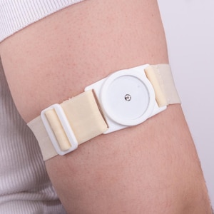Op de afbeelding: Een wit medisch apparaat met een ronde sensor bevestigd aan een beige stoffen band. De band wordt op de bovenarm gedragen.
