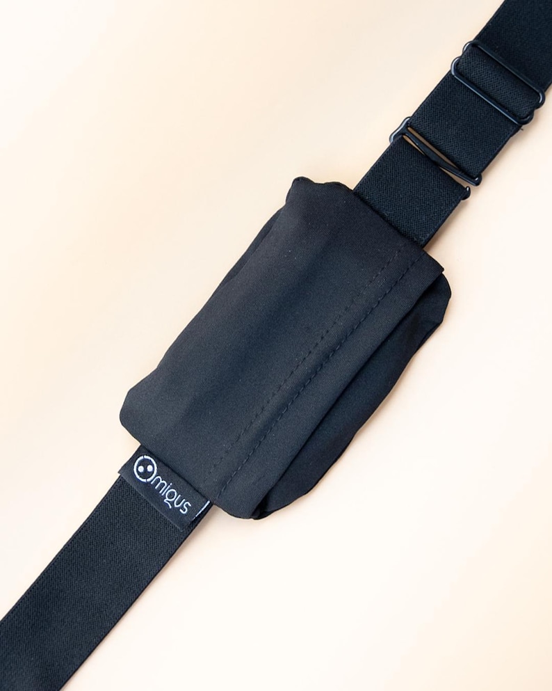May include: Black adjustable strap with a small pouch. The strap has a metal buckle and a small tag with the brand name "@migus". The pouch is made of a soft, dark fabric.