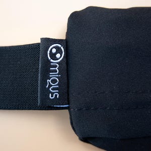 May include: Close-up of a black sleep mask with a black elastic band. A small black tag with the white embroidered brand name "miq&uuml;s" and a smiling face logo is attached to the mask.