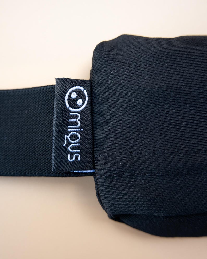 May include: Close-up of a black sleep mask with a black elastic band. A small black tag with the white embroidered brand name "miq&uuml;s" and a smiling face logo is attached to the mask.